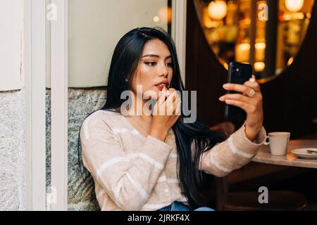 Junge ethnische Frauen, die Lippenstift auf die Lippen auftragen, während sie auf den Handybildschirm auf dem Fensterbrett der Cafeteria schauen Stockfoto
