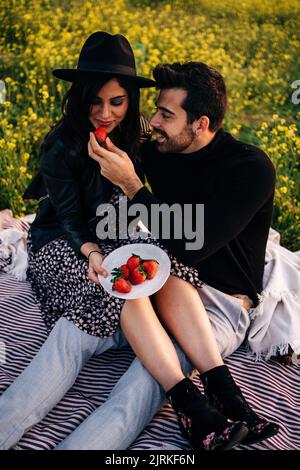 Junge zufriedene ethnische Männer füttern Freundin mit reifen Erdbeeren, während sie auf Stoff gegen blühende Blumen sitzen Stockfoto