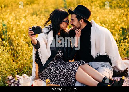 Junge fröhliche ethnische Frau mit Freund, die auf der Fotokamera Selbstporträt macht, während sie auf der Wiese sitzt Stockfoto