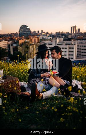 Junge zufriedene ethnische Männer füttern Freundin mit reifen Erdbeeren, während sie auf Stoff gegen blühende Blumen sitzen Stockfoto