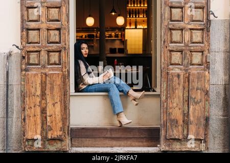 Junge ethnische Frauen in legerer Kleidung, die sich auf dem Fensterbrett der Cafeteria Notizen machen Stockfoto