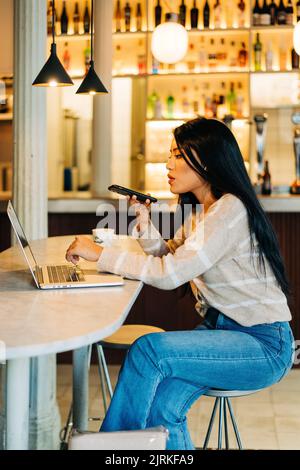 Seitenansicht einer jungen ethnischen Frau, die am Cafeteria-Tisch mit Netbook eine Sprachnachricht auf dem Handy sendet Stockfoto