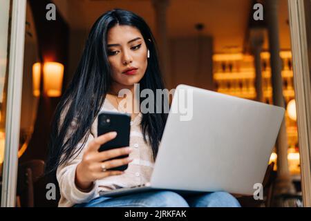 Ein niedriger Winkel junger asiatischer Frauen, die sich mit einem Handy unterhalten, während sie mit einem Netbook in der Cafeteria sitzen Stockfoto