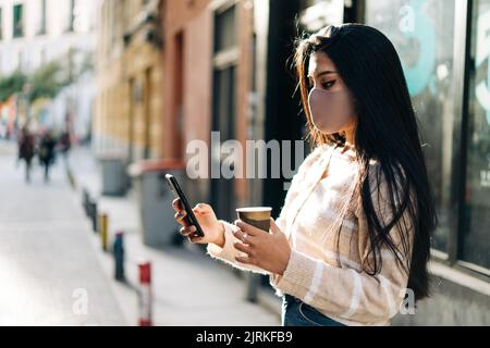 Seitenansicht der ethnischen Frau in Stoff Gesichtsmaske mit heißem Getränk zum Surfen im Internet auf Handy in der Stadt zu gehen Stockfoto