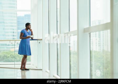 Eine ziemlich nachdenkliche Geschäftsfrau mit digitalem Tisch, die durch das Fenster in ihrem Büro schaut Stockfoto