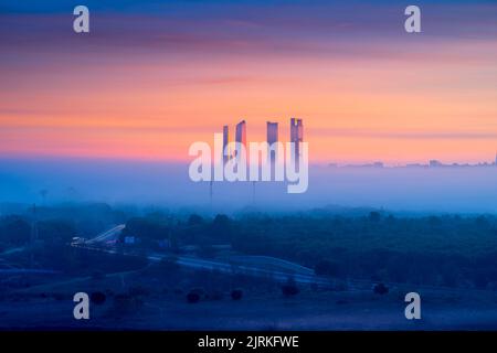Luftaufnahme der Stadtlandschaft mit einem Komplex von modernen Wolkenkratzern, die bei Sonnenaufgang über dem nebligen Madrid aufragen, mit bunt bewölktem Himmel Stockfoto
