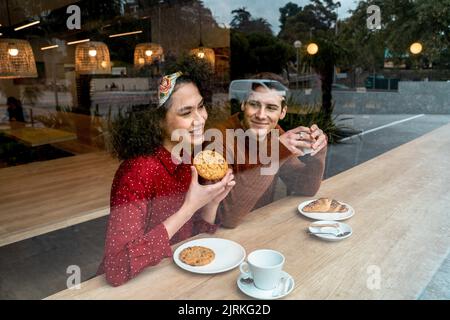 Durch ein Glas fröhliches junges, verliebtes multirassisches Paar, das an der Holztheke sitzt und in der gemütlichen Cafeteria leckeres Gebäck und Kaffee genießt Stockfoto