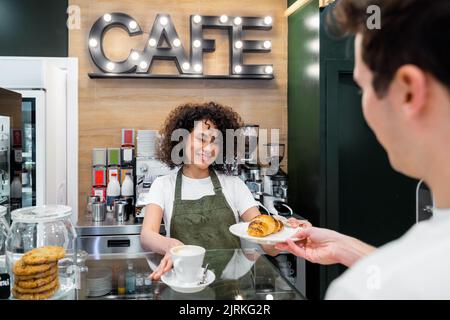 Die freundliche, ethnische Barista serviert dem Gast eine Tasse Cappuccino und ein frisches, süßes Croissant, während sie an der Theke im Café steht Stockfoto