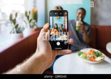 Crop unkenntlich männlich fotografieren lächelnde multiethnische Partner auf Handy gegen den Tisch mit Essen im Restaurant Stockfoto