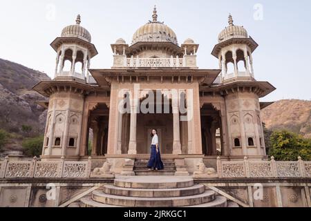 Seitenansicht einer fröhlichen asiatischen Frau, die lächelt und die Kamera anschaut, während sie in der Nähe des gealterten Gaitor Ki Chhatriyan-Gebäudes in Jaipur, Indien, steht Stockfoto