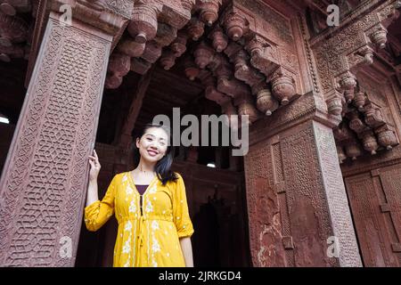 Glückliche, ethnische Asiatin, die in gelbem Kleid in einem veralterten, gewölbten Agra Fort in Uttar Pradesh, Indien, die Kamera anschaut Stockfoto