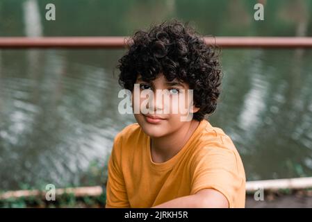 Fröhlicher Junge mit dunklem lockigem Haar in legerer Kleidung, der in der Nähe des ruhigen Flusses sitzt und tagsüber die Kamera anschaut Stockfoto