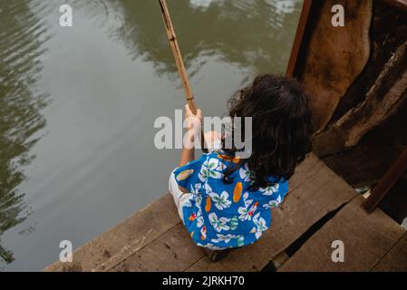 Von oben Rückansicht eines ruhigen, nicht erkennbaren Jungen in legerer Kleidung, der auf einem hölzernen Pier in der Nähe des Teichs sitzt und tagsüber angeln kann Stockfoto