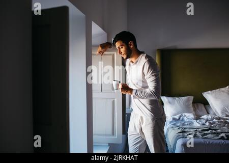 Seitenansicht eines ruhigen jungen Mannes in weißer Schlafkleidung, der aus dem Fenster schaute, während er sich auf die Fensterläden lehnte und Kaffee im Schlafzimmer trank Stockfoto