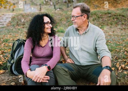 Fröhliches reifes Paar in legerer Kleidung, das sich auf dem Boden im Herbstpark anschaut Stockfoto