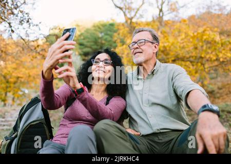 Inhalt Seniorenpaar in legerer Kleidung und Brille beim Selfie auf dem modernen Smartphone, während sie auf Gras im Herbstpark sitzen Stockfoto