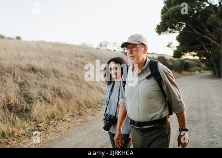 Fröhliches reifes Paar in legerer Kleidung und professionelle Fotografie Kamera zu Fuß auf dem Weg in der Nähe von Wiese in der Landschaft Stockfoto