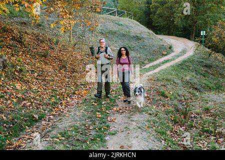 Ganzkörper-positive reife Paare tragen lässige Outfits, die zusammen mit dem liebenswerten australischen Schäferhund in einem sonnigen Herbstpark spazieren gehen Stockfoto