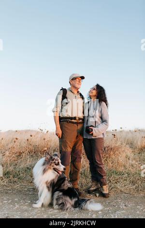 Ganzkörper-fröhliches reifes Paar in lässiger Kleidung, umarmt und stehend auf einer Wiese auf dem Land in der Nähe des entzückenden australischen Schäferhundes Stockfoto