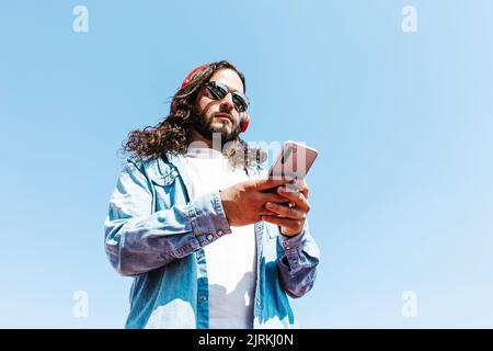 Von unten bärtiger lateinamerikanischer Mann mit moderner Sonnenbrille, der am sonnigen Tag auf dem Handy und kabellosen Kopfhörern auf blauem Hintergrund surft Stockfoto