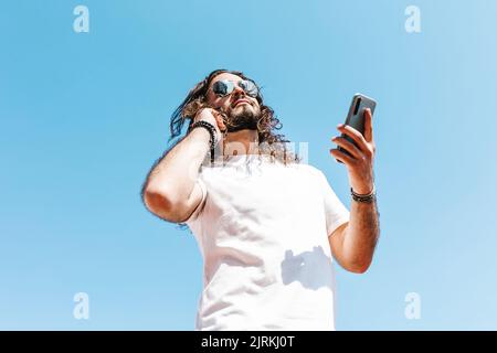 Von unten der bärtige lateinamerikanische Mann in weißem T-Shirt und moderne Sonnenbrille mit Handy auf blauem Hintergrund an sonnigen Tag Stockfoto