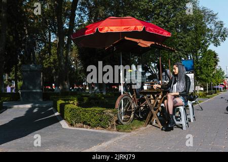 Junge lateinamerikanische Hündin mit Afro-Zöpfen, die mit gekreuzten Beinen am Tisch unter Sonnenschutz im sonnigen Park sitzt Stockfoto