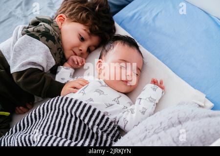 Von oben ein charmanter kleiner Junge, der kleine Geschwister umarmt und in einem gemütlichen Bett friedlich zusammenschläft Stockfoto