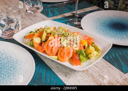 Köstlicher Salat mit frisch geschnittenen Tomaten und Avocado mit mariniertem Lachs unter Sprossen neben einem Glas Rotwein auf dem Tisch Stockfoto