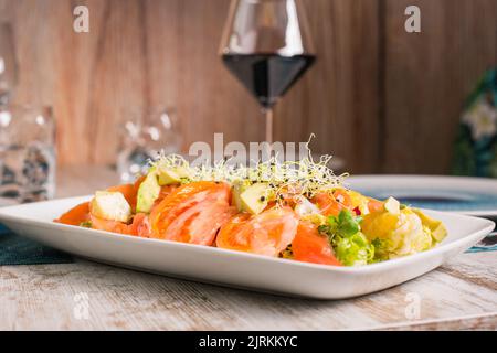 Köstlicher Salat mit frisch geschnittenen Tomaten und Avocado mit mariniertem Lachs unter Sprossen neben einem Glas Rotwein auf dem Tisch Stockfoto