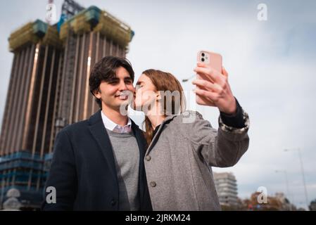 Glückliche junge Frau umarmt und küsst Freund, während Selfie auf dem Smartphone stehend zusammen auf der Straße der Stadt Stockfoto