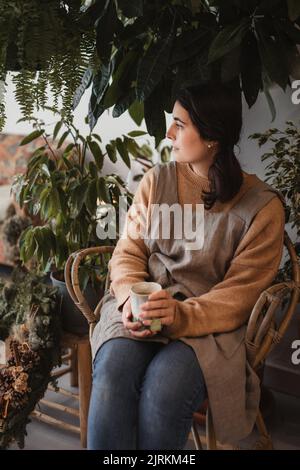 Nachdenkliche, entspannte Frau in legerer Kleidung, die wegschaut und kontempliert, während sie auf dem Sofa neben einem kleinen Holztisch sitzt und leckeren Kaffee-Agai trinkt Stockfoto