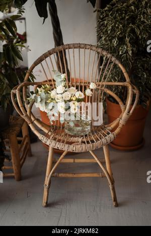 Hoher Winkel der schönen Zusammensetzung von frischen weißen Blumen und grünen Laub in Glas transparente Vase stehen auf Vintage braun Korbstuhl gegen Stockfoto