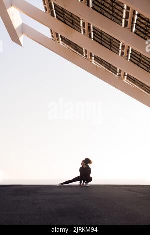 Afroamerikanische Athletin, die beim Training unter einem modernen Haus Seitenhock ausführt, während sie in die Stadt blickt Stockfoto