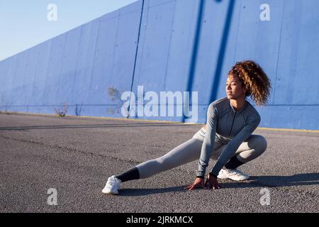 Erwachsene Sportlerin in Sportbekleidung beim Hocken während des Trainings auf asphaltierter Straße mit Schatten in der Stadt Stockfoto