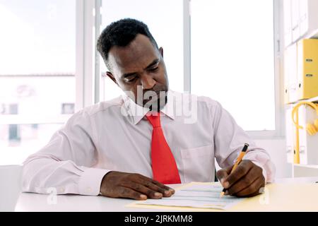 Reifer afroamerikanischer männlicher Manager in weißem Hemd mit Krawatte, der auf einem Papierdokument am Tisch im Arbeitsbereich notiert Stockfoto