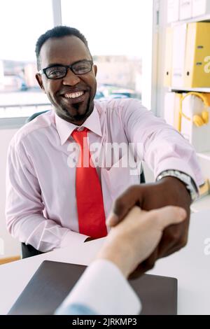 Lächelnder afroamerikanischer männlicher Chef in formellem Outfit und Brillen, die die Hand eines anonymen Kollegen aus der Ernte schüttelten, während sie die Kamera betraten Stockfoto