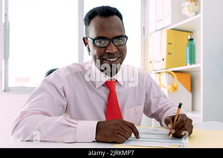 Reifer afroamerikanischer Manager mit Ohrstöpsel in weißem Hemd mit Krawatte und Brille, der auf einem Papierdokument am Tisch im Arbeitsbereich Notiz nimmt Stockfoto