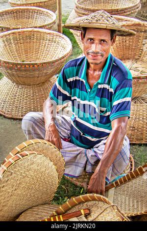 Narayanganj, Dhaka, Bangladesch. 25. August 2022. Ein Verkäufer sitzt inmitten von Kunsthandwerk, die auf einem Wochenmarkt in Narayanganj, Bangladesch, ordentlich zum Verkauf angeboten werden. Die Körbe, Handfächer, Fischfangfallen, Caps und Handtaschen spiegeln die Kultur, das Erbe und den kreativen Ausdruck von Menschen mit einzigartigen künstlerischen Fähigkeiten wider. Bangladesch verdient jährlich rund 20 Millionen US-Dollar aus dem Export von Handwerksprodukten. Die Facharbeiter haben ihr Handwerk über viele Jahre verfeinert. Hergestellt aus Holz und Bambus, zeichnen sich die Artikel durch die Zweckmäßigkeit, Nachhaltigkeit und Umweltfreundlichkeit aus Stockfoto