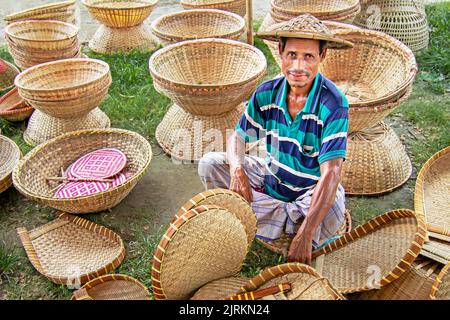 Narayanganj, Dhaka, Bangladesch. 25. August 2022. Ein Verkäufer sitzt inmitten von Kunsthandwerk, die auf einem Wochenmarkt in Narayanganj, Bangladesch, ordentlich zum Verkauf angeboten werden. Die Körbe, Handfächer, Fischfangfallen, Caps und Handtaschen spiegeln die Kultur, das Erbe und den kreativen Ausdruck von Menschen mit einzigartigen künstlerischen Fähigkeiten wider. Bangladesch verdient jährlich rund 20 Millionen US-Dollar aus dem Export von Handwerksprodukten. Die Facharbeiter haben ihr Handwerk über viele Jahre verfeinert. Hergestellt aus Holz und Bambus, zeichnen sich die Artikel durch die Zweckmäßigkeit, Nachhaltigkeit und Umweltfreundlichkeit aus Stockfoto