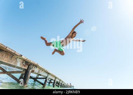 Von unten ein fröhlicher Teenager im Moment, in dem er vom Pier im Meer springt und das Sommerwochenende an einem sonnigen Tag genießt