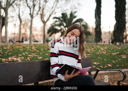 Fröhliche junge Studentin, die mit offenem Buch in den Händen auf der Bank sitzt, während sie den Herbsttag im Park verbringt Stockfoto