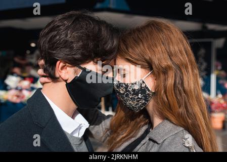 Seitenansicht eines glücklichen jungen verliebten Paares, das Schutzmasken trägt, die nahe stehen und umarmen, während es ein romantisches Date mit geschlossenen Augen hat Stockfoto