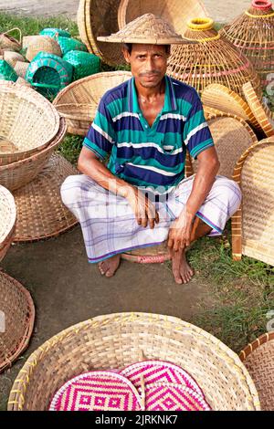 Narayanganj, Dhaka, Bangladesch. 25. August 2022. Ein Verkäufer sitzt inmitten von Kunsthandwerk, die auf einem Wochenmarkt in Narayanganj, Bangladesch, ordentlich zum Verkauf angeboten werden. Die Körbe, Handfächer, Fischfangfallen, Caps und Handtaschen spiegeln die Kultur, das Erbe und den kreativen Ausdruck von Menschen mit einzigartigen künstlerischen Fähigkeiten wider. Bangladesch verdient jährlich rund 20 Millionen US-Dollar aus dem Export von Handwerksprodukten. Die Facharbeiter haben ihr Handwerk über viele Jahre verfeinert. Hergestellt aus Holz und Bambus, zeichnen sich die Artikel durch die Zweckmäßigkeit, Nachhaltigkeit und Umweltfreundlichkeit aus Stockfoto