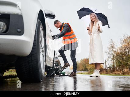 Schöne junge Frau mit Regenschirm, Daumen hoch und lächelnd, während Pannenhelfer Reifen mit Luftkompressor aufbläst. Auto Mechaniker Reparatur Auto, während Frau zeigt Genehmigung Geste. Stockfoto