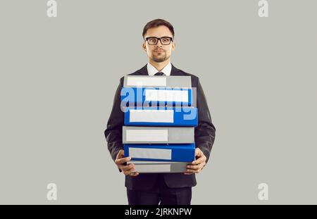 Angespannter männlicher Büroangestellter mit gestresster Expression, der Stapel schwerer Ordner auf grauem Hintergrund hält. Stockfoto