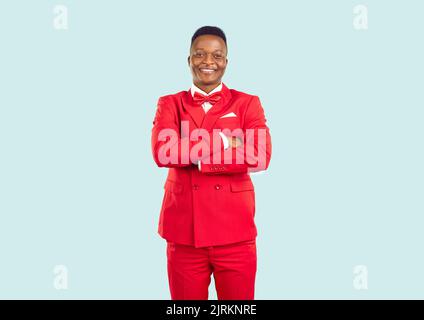 Fröhlicher, hübscher junger Mann in rotem Anzug, der im Modestudio mit gefalteten Armen steht Stockfoto
