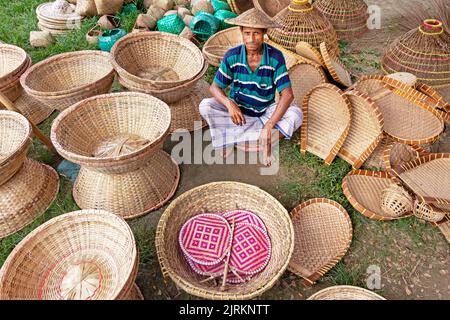 Narayanganj, Dhaka, Bangladesch. 25. August 2022. Ein Verkäufer sitzt inmitten von Kunsthandwerk, die auf einem Wochenmarkt in Narayanganj, Bangladesch, ordentlich zum Verkauf angeboten werden. Die Körbe, Handfächer, Fischfangfallen, Caps und Handtaschen spiegeln die Kultur, das Erbe und den kreativen Ausdruck von Menschen mit einzigartigen künstlerischen Fähigkeiten wider. Bangladesch verdient jährlich rund 20 Millionen US-Dollar aus dem Export von Handwerksprodukten. Die Facharbeiter haben ihr Handwerk über viele Jahre verfeinert. Hergestellt aus Holz und Bambus, zeichnen sich die Artikel durch die Zweckmäßigkeit, Nachhaltigkeit und Umweltfreundlichkeit aus Stockfoto