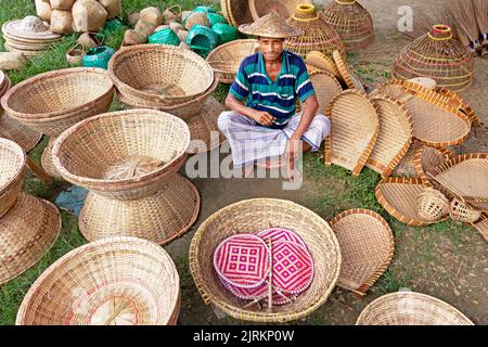 Narayanganj, Dhaka, Bangladesch. 25. August 2022. Ein Verkäufer sitzt inmitten von Kunsthandwerk, die auf einem Wochenmarkt in Narayanganj, Bangladesch, ordentlich zum Verkauf angeboten werden. Die Körbe, Handfächer, Fischfangfallen, Caps und Handtaschen spiegeln die Kultur, das Erbe und den kreativen Ausdruck von Menschen mit einzigartigen künstlerischen Fähigkeiten wider. Bangladesch verdient jährlich rund 20 Millionen US-Dollar aus dem Export von Handwerksprodukten. Die Facharbeiter haben ihr Handwerk über viele Jahre verfeinert. Hergestellt aus Holz und Bambus, zeichnen sich die Artikel durch die Zweckmäßigkeit, Nachhaltigkeit und Umweltfreundlichkeit aus Stockfoto