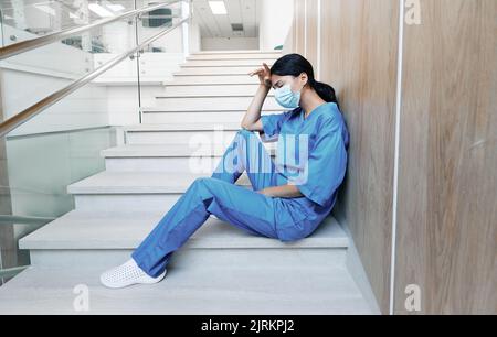 Müde Ärztin nach hartem Arbeitstag in der medizinischen Klinik auf Stufen im Krankenhaus sitzend mit geschlossenen Augen und medizinischer Maske. Pandemia-Coronavirus Stockfoto