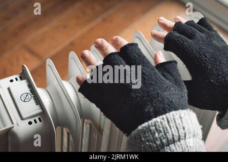 Nahaufnahme der Hände einer Person, die eine Wolljacke und schwarze wollige fingerlose Handschuhe in der Nähe eines Heizkörpers trägt, der versucht, sich im Winter warm zu halten Stockfoto